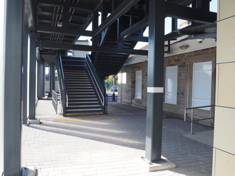 Kilwinning new footbridge: This picture shows the area between the old station building and the new glazed screen in front of it, with the stairs down from the new footbridge. Lift out of shot to the right. Photo of Kilwinning new footbridge
